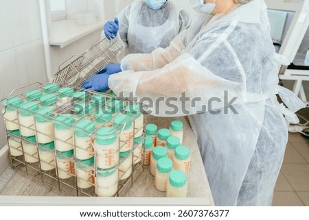 Two lab workers in protective wear handling donor breast milk containers and organizing them into metal baskets for further processing in milk bank environment.