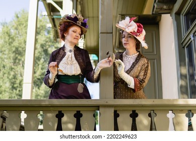 Two Ladies In Early 20th Century Costumes Are Talking On The Manor Terrace On A Summer Day. Women Are Considering A Novelty - Sunglasses
