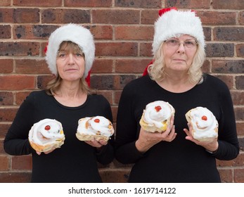 Two Ladies Are Holding Iced Buns  Pretending They Are Breasts With Cherries As Nipples And Wear Christmas Festive Hats As A Joke.They Have Serious Faces And Do Not Look Amused In Contrast.Humour.Image