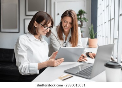 Two lady colleagues discussing new project, businesswomen using digital tablet together in office. Happy business colleagues in coworking space using tablet - Powered by Shutterstock