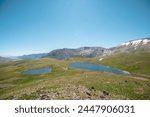 Two lakes, sunlit green meadow, alpine valley and mountain range. Blue sky gradient reflected in water. Top view from rocks to dirt road between two beautiful lakes in high mountains in sunny day.