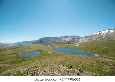 Two lakes, sunlit green meadow, alpine valley and mountain range. Blue sky gradient reflected in water. Top view from rocks to dirt road between two beautiful lakes in high mountains in sunny day. - Powered by Shutterstock