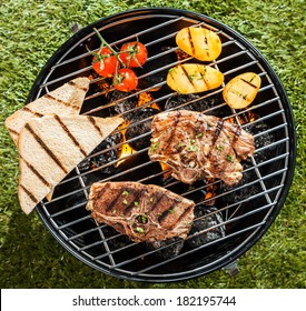 Two Lamb Chops Cooking On A BBQ On A Portable Grill With Tomato, Potatoes And Slices Of Toast On A Sunny Summer Day Outdoors On The Grass, Overhead View