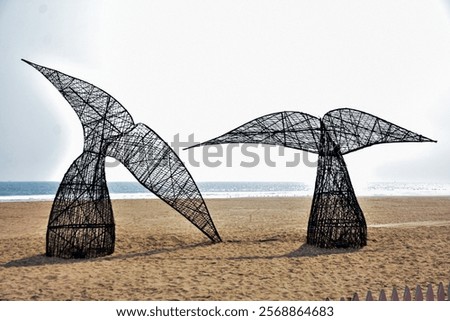 Two large, abstract whale tail sculptures made of dark woven material rise from a sandy beach, contrasting with the calm ocean and distant horizon.