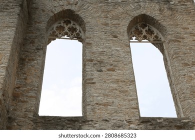 Two large, arched window openings built into a weathered, ancient stone wall, likely belonging to a ruined medieval church or monastery. The tops of the windows are decorated with intricate, crumbling Gothic stone tracery. The window panes are entirely missing, framing a clear, pale overcast sky through the open masonry structure.