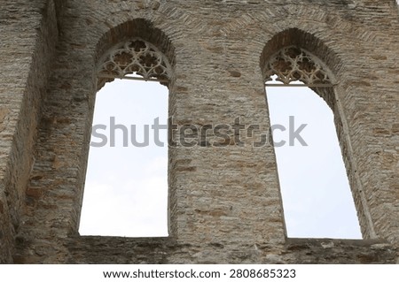 Two large, arched window openings built into a weathered, ancient stone wall, likely belonging to a ruined medieval church or monastery. The tops of the windows are decorated with intricate, crumbling Gothic stone tracery. The window panes are entirely missing, framing a clear, pale overcast sky through the open masonry structure.