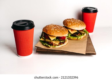 Two Large Burgers On Kraft Paper And A Wooden Board, With Two Red Paper Cups Of Coffee On A White Background, A Wooden Table. Fast Junk American Food. Large Portions. Takeaway Delivery. Vegetarian.