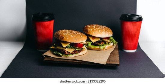 Two Large Burgers On Kraft Paper And A Wooden Board, With Two Red Paper Cups Of Coffee On A Grey Paper, White Background, A Wooden Table. Fast Junk American Food. Takeaway Delivery. Vegetarian.