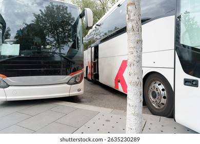 Two large buses positioned side by side at a busy bus terminal during daytime. The area features greenery and a modern urban setting.