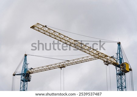 Two large construction cranes are actively positioned at a construction site, lifting materials as workers prepare the framework for a new house. The sky above is overcast