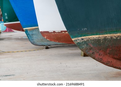 Two large fishing vessels on drydock in storage. One boat is blue with the number five in white.The second is green with the white lettering numbers of eight, ten and twelve. Both boats have red hulls