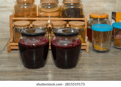 Two large jars filled with deep purple jam are beautifully displayed on a rustic wooden table. Behind them, a charming wooden rack holds various spices and jars, creating a warm kitchen ambiance.
