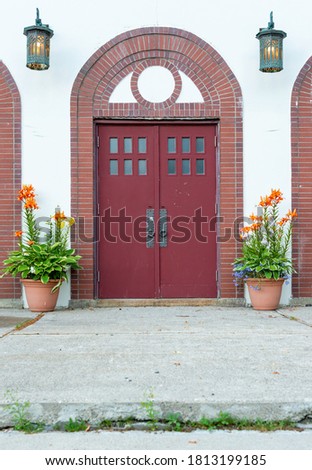 Two large red, double doors with six small windows and a round metal door handle in each door. The exterior of the building has white stucco with vintage brass lanterns and two clay flower pots.