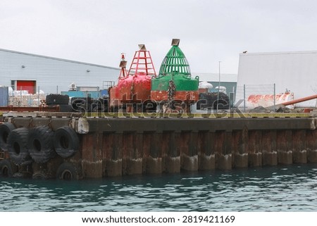Two large red and green navigation buoys sit on a weathered industrial dock at a busy harbor, surrounded by tires, shipping containers and rusted metal, conveying maritime industry and coastal work