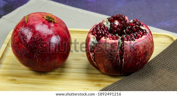 Two large large red pomegranate fruits. Whole red\
ripe pomegranate. Pomegranate fruit open which is divided into five\
parts which are held together on wooden tray on blue background.\
Banner.