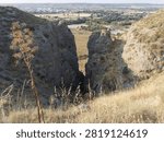 Two large rock formations stand on either side of a narrow canyon. A dry grass field is visible below, with distant hills and farmland. Sunlight brightens the scene.