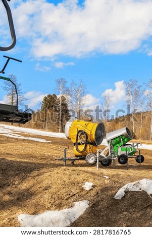 Two large snowmaking machines stand on a ski slope in a forested area