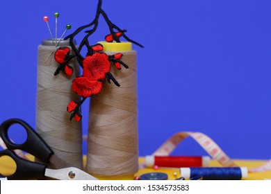 Two large spools of thread stand on a blue background. In the photo there is an application in the form of red flowers, scissors for sewing, buttons and tape measure. Needlework concept.