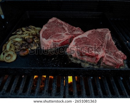 Two large, thick beef T-bone steaks are grilling on a cast-iron grate under a barbecue flame. There are white onions cut into rings, fried in butter. The beefsteaks are organic, grass-fed beef, and thick.