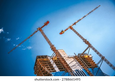 Two large tower cranes are actively engaged in the construction of a high-rise building, reaching towards the clear sky