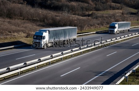 Two large trucks with gray trailers are driving on a highway through a rural area. Winter period without snow. Brown color prevails in the forest above the road.

