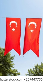 Two large, vertical Turkish flags hang side-by-side from a wire against a bright blue sky, partially framed by the green foliage of trees below.