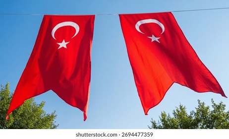 Two large, vertical Turkish flags hang side-by-side from a wire against a bright blue sky, partially framed by the green foliage of trees below.
