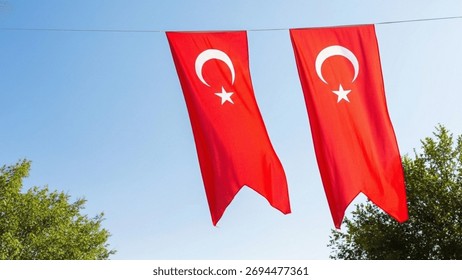 Two large, vertical Turkish flags hang side-by-side from a wire against a bright blue sky, partially framed by the green foliage of trees below.
