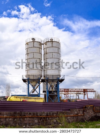 Two large white cement silos stand against a blue sky with clouds. Blue metal supports hold silos above a low purple fence