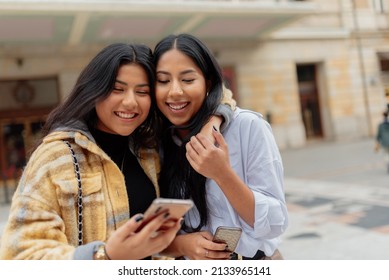 Two Latin Women Looking At Each Other And Holding Their Mobile Phones In The Street. Hispanic Sister Having A Good Time Together Sightseeing.