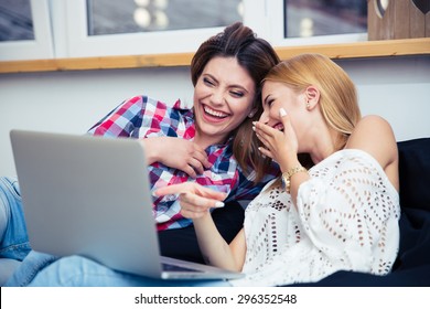 Two laughing girls sitting on sofa and watching movie on laptop