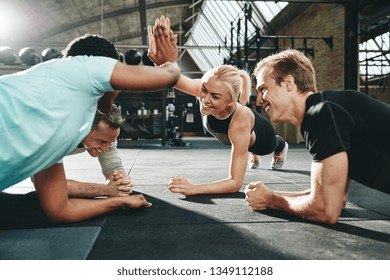 Two Laughing Young Women In Sportswear High Fiving Each Other During A Planking Class At The Gym With A Couple Of Male Friends