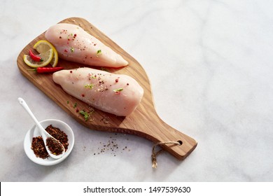 Two Lean Healthy Low Fat Raw Chicken Breasts Sprinkled With Spices And Chili On A Wooden Cutting Board On A White Marble Kitchen Counter With Copy Space In An Overhead View