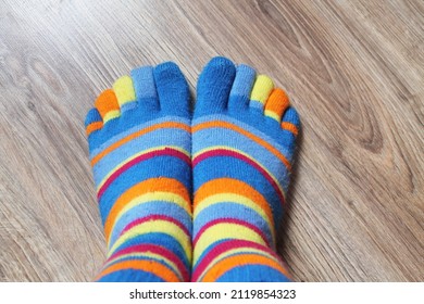 Two legs stand together on a brown laminate, close-up, top view. On the feet are socks in multi-colored stripes. Socks are worn on each finger.