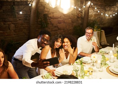 Two Lesbian Brides Boasting With Their Wedding Rings By Served Festive Table While Looking At Smartphone Screen Held By Young Black Man