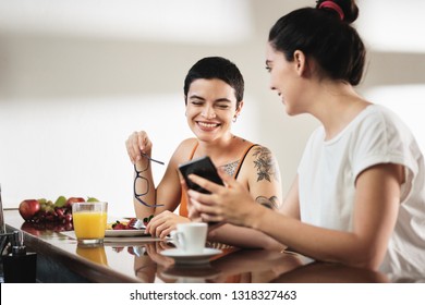 Two lesbians at home, girl eating fruit and food for snack, partner showing funny video on mobile telephone. Happy homosexual people domestic lifestyle. - Powered by Shutterstock