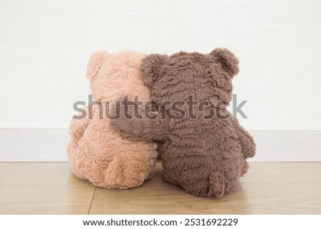 Two light and dark brown soft teddy bears hugging each other and sitting on wooden floor at light gray wall background. Closeup. Back view.