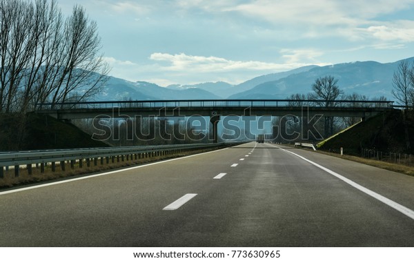 Two line wide highway on a cloudy\
winter day leading to the mountains through rural\
landscape