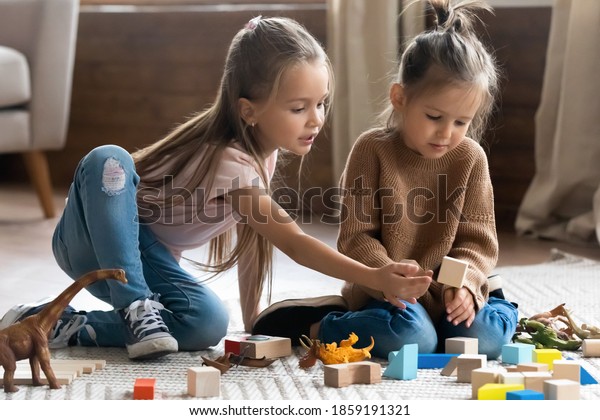Two little 6s 7s sisters pretty daughters spend free playtime seated on carpet warm floor in modern living room play together wooden brick and dinosaurs toys set. Friendship, fun and leisure concept