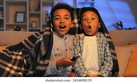 Two little African American brothers are sitting on the sofa covered with a blanket and watching TV. Two multiracial boys get scared while watching a horror movie. - Powered by Shutterstock