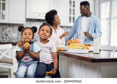 Two Little African American Girls Sisters With Cookies, Hugging On Bright Kitchen While Their Affectionate Lovely Parents Talking On Blur Background. Happy Family Enjoying Morning Time At Home.