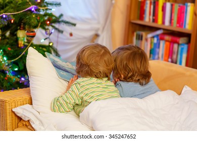 Two Little Blond Children Reading A Book Together In Bed Near Christmas Tree With Lights And Illumination. Happy Family, Two Friends, Kid Twins Boys.