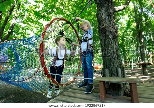 Two\
little boy in climbing gear is walking along a rope road in an\
adventure Park, holding onto a rope and a\
carabiner.