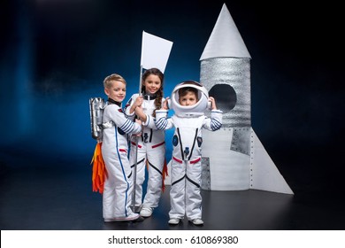 Two little boys and girl in space suits holding flag near rocket  and smiling at camera