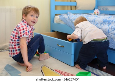 Two little brothers play toys near bed with big drawer in room 