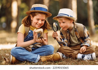 Two little children boy and girl with backpacks looking examining plants and insects in a jar through magnifying glass while exploring forest nature and environment on sunny day during outdoor lesson - Powered by Shutterstock