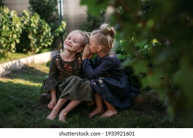 Two little cute girls are sitting in the garden and sharing secrets. One girl whispers something in another's ear.