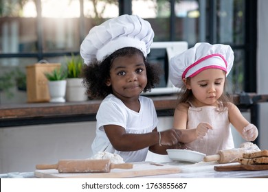 Two Little Girl Friends With Different Skin Types Are Enjoying Learning How To Make Bread In Kitchen, Bias Racial Concept