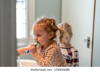 Two Little Girls In The Bathroom Taking Turns Brushing Teeth In Morning Sunlight