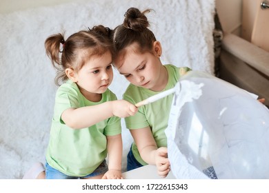 Two Little Girls With Dark Hair And Light Skin In Light Green Make A Paper Mache Craft